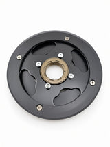 A circular black metal mechanical component with a central gold-toned ring, six silver screws, and cutout slots, photographed on a plain white background.