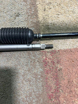 Steering Gear Assembly for the Coleman KT196 196cc 6.5 Hp Go-Kart (Blemished) featuring a close-up of metal components, including a steering shaft, tie rods, and a steering gearbox on a grey surface.